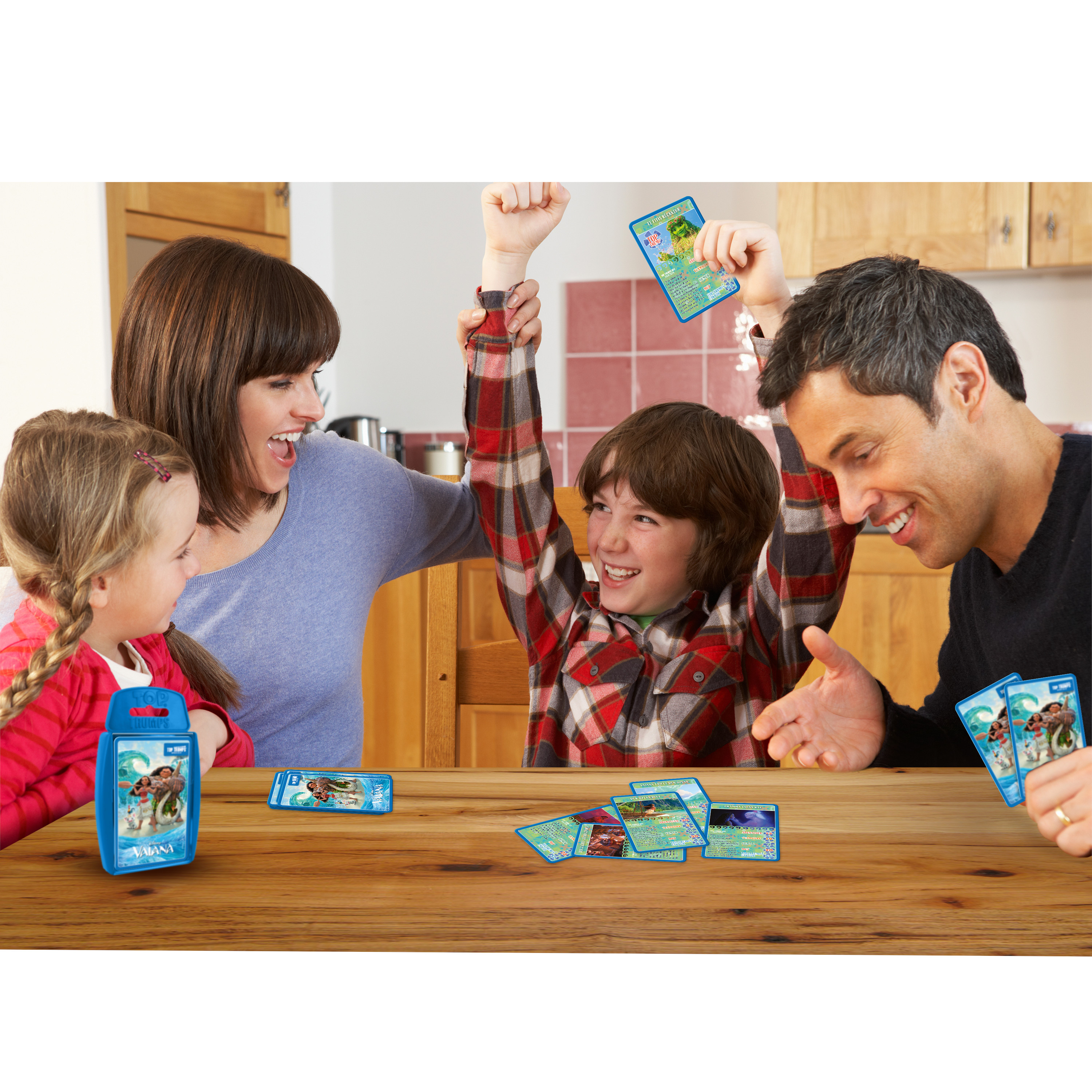 Eine Familie spielt ein Kartenspiel, wobei ein Kind, das gewinnt, die Arme hochreißt. Karten liegen auf dem Tisch.