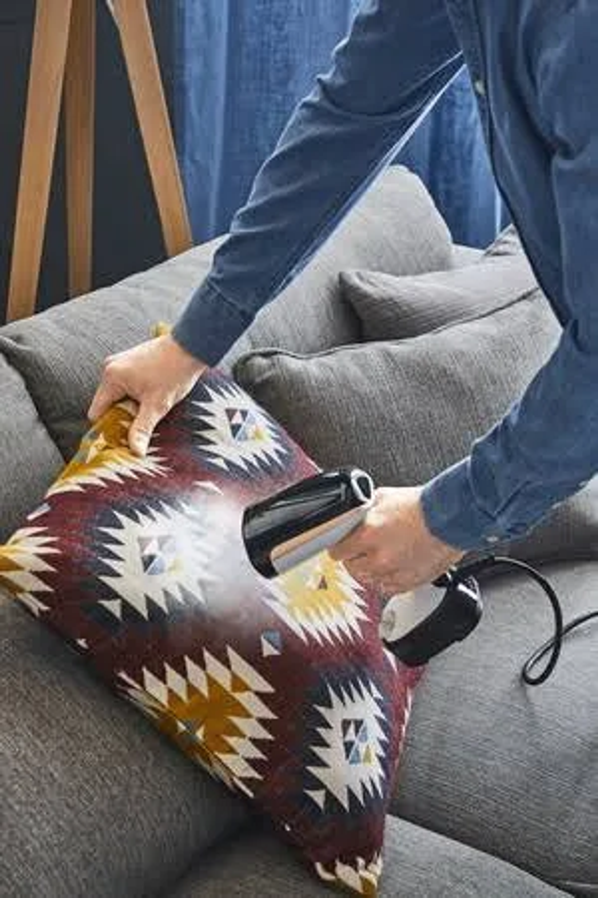 Persona che stira un cuscino a fantasia su un divano grigio con un vaporizzatore portatile.