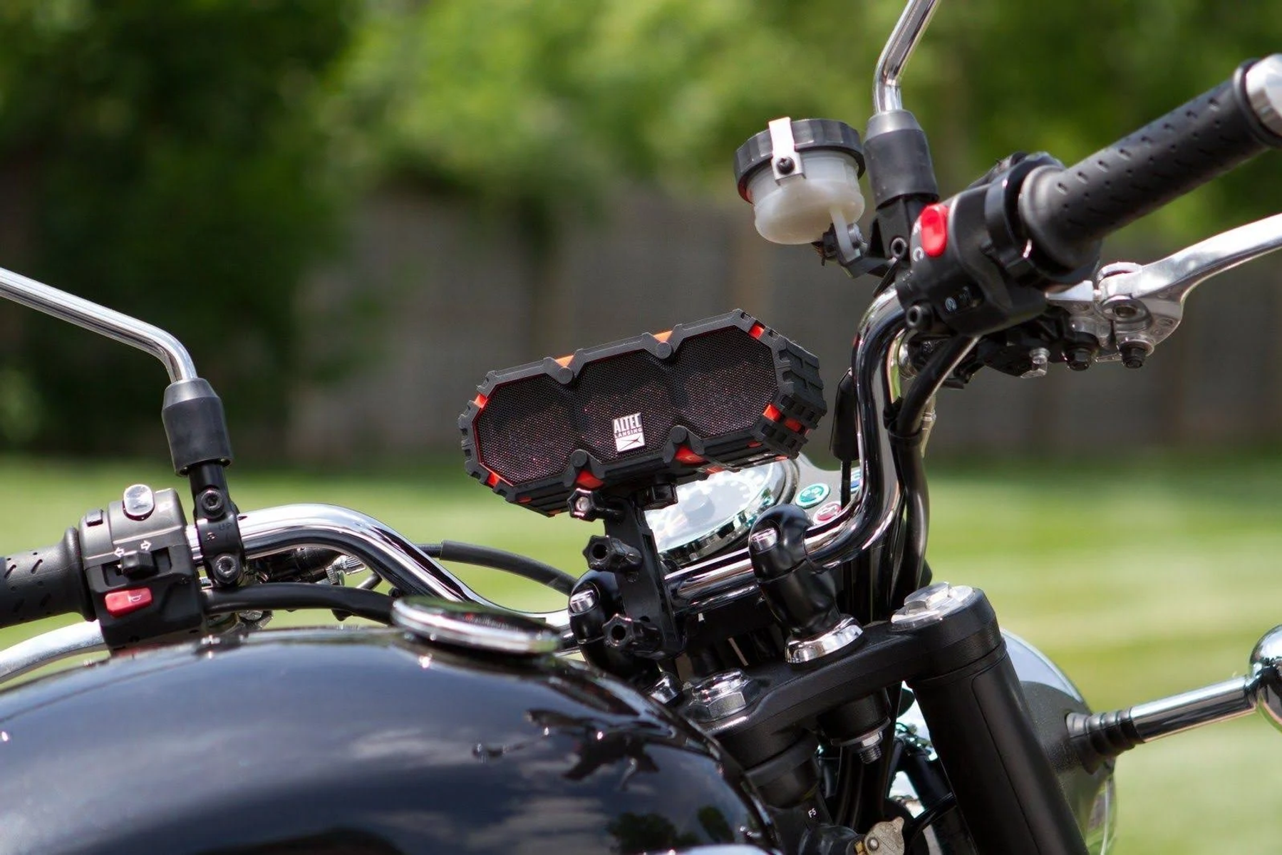 Czarny i pomarańczowy głośnik zamontowany na kierownicy motocykla, rozmyte zielone tło.