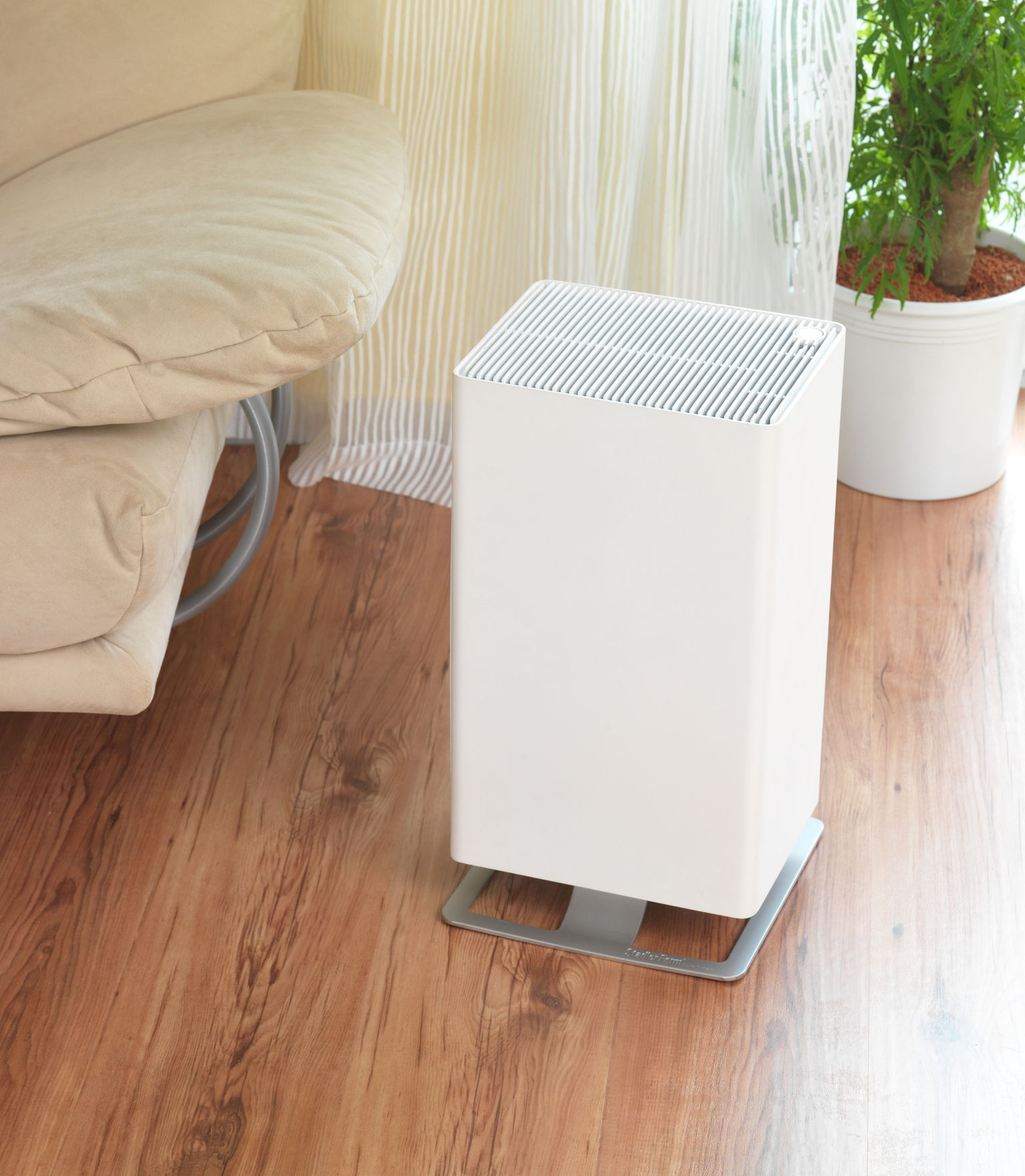 White air purifier on a silver stand, near a sofa and a plant in a room with wooden floor.