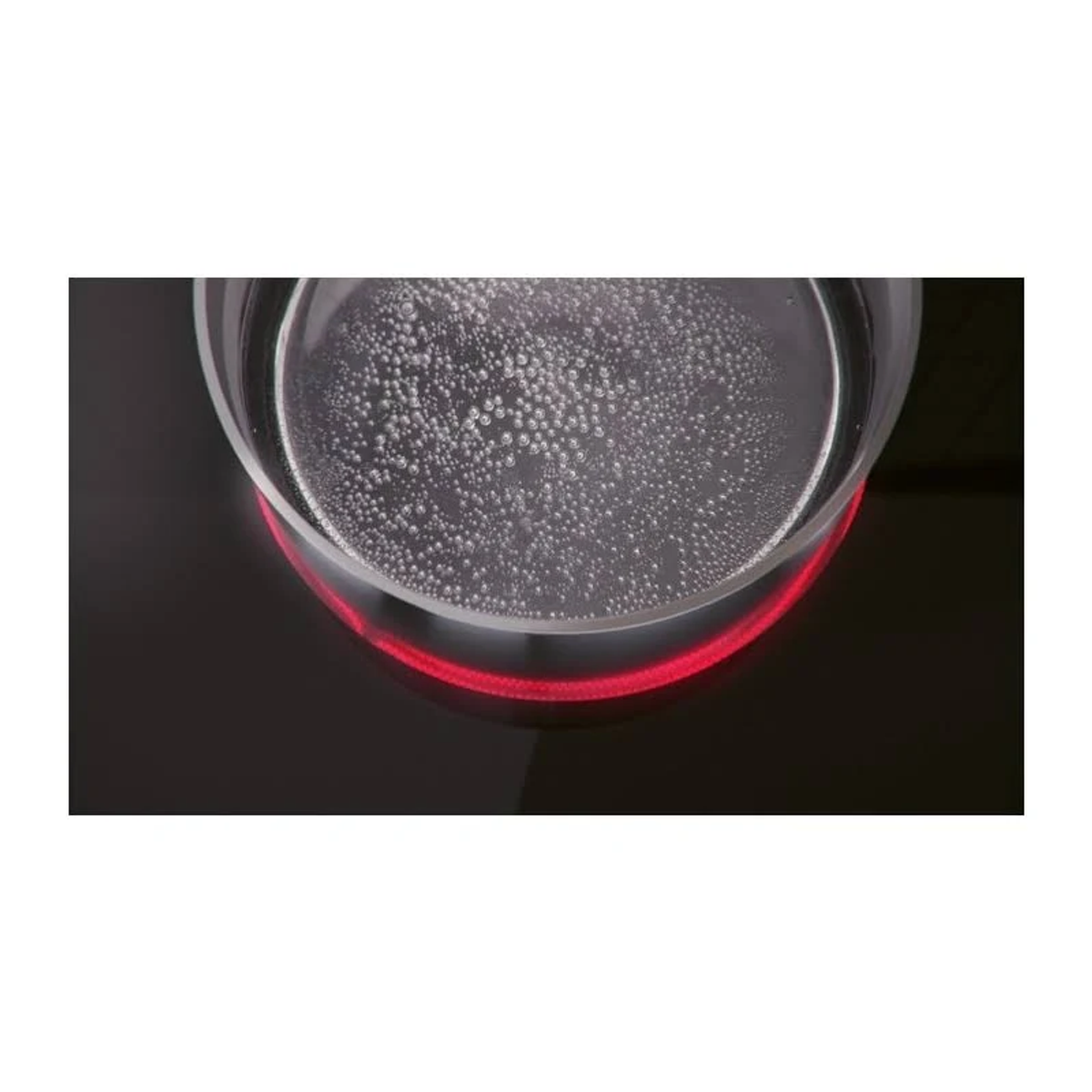 Zbliżenie szklanej miski na czarnej płycie grzejnej. Woda w środku ma bąbelki. Widoczne jest czerwone światło.