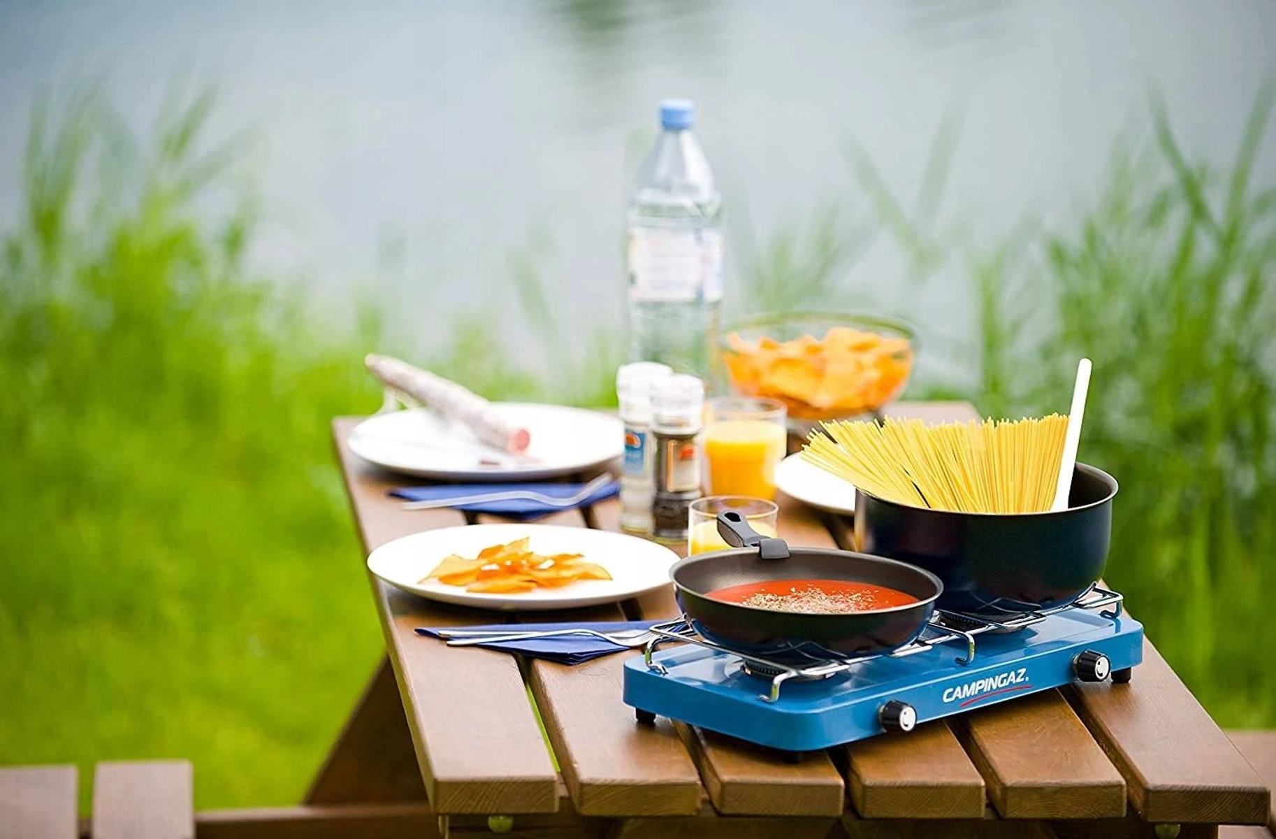 Un tavolo da picnic con cibo e attrezzature da cucina vicino all'acqua e all'erba verde nella natura.