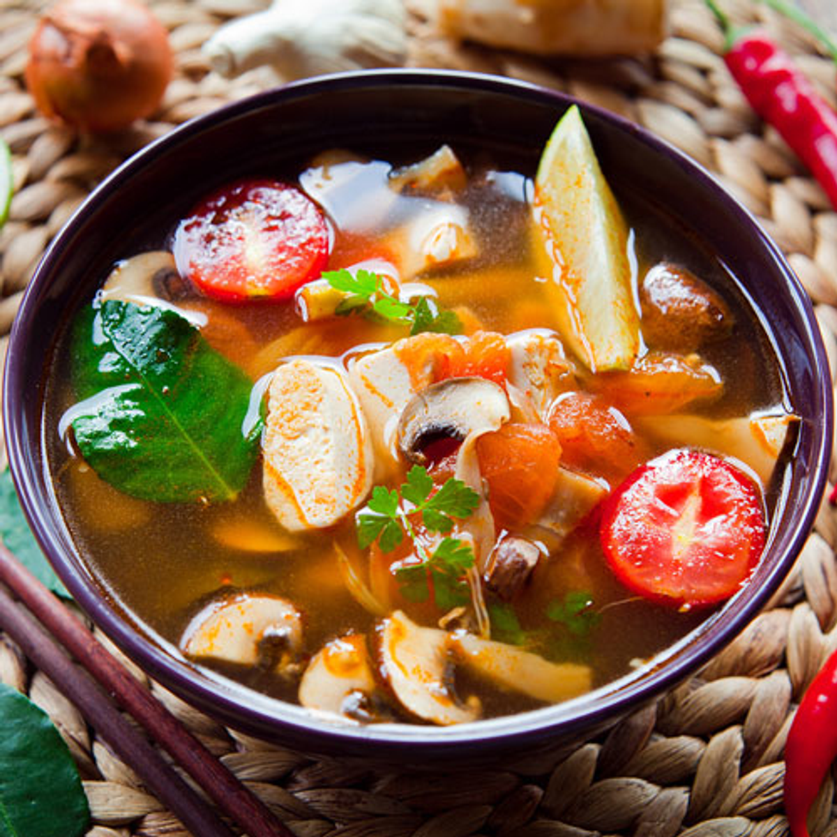 Eine Schüssel mit thailändischer Suppe mit Tomaten, Pilzen und Limette. Essstäbchen sind zu sehen.