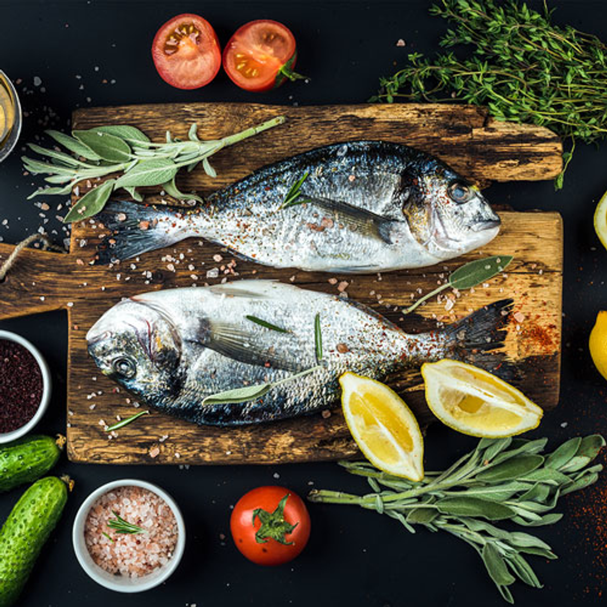 Zwei Fische auf Holzbrett mit Kräutern, Tomaten, Zitronenscheiben und Gewürzen.