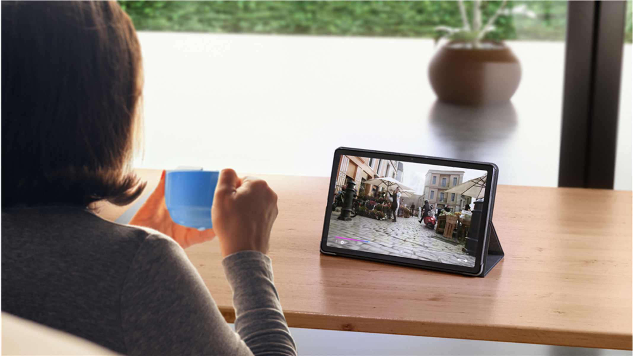 Osoba oglądająca wideo na tablecie, trzymając niebieski kubek, siedząca przy oknie.
