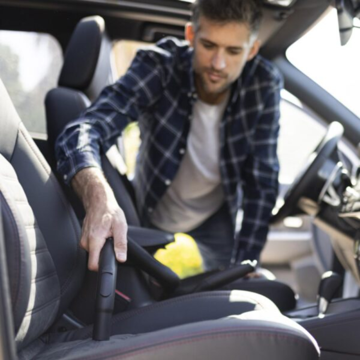 Hombre limpiando un asiento de coche con una aspiradora. Lleva una camisa a cuadros.