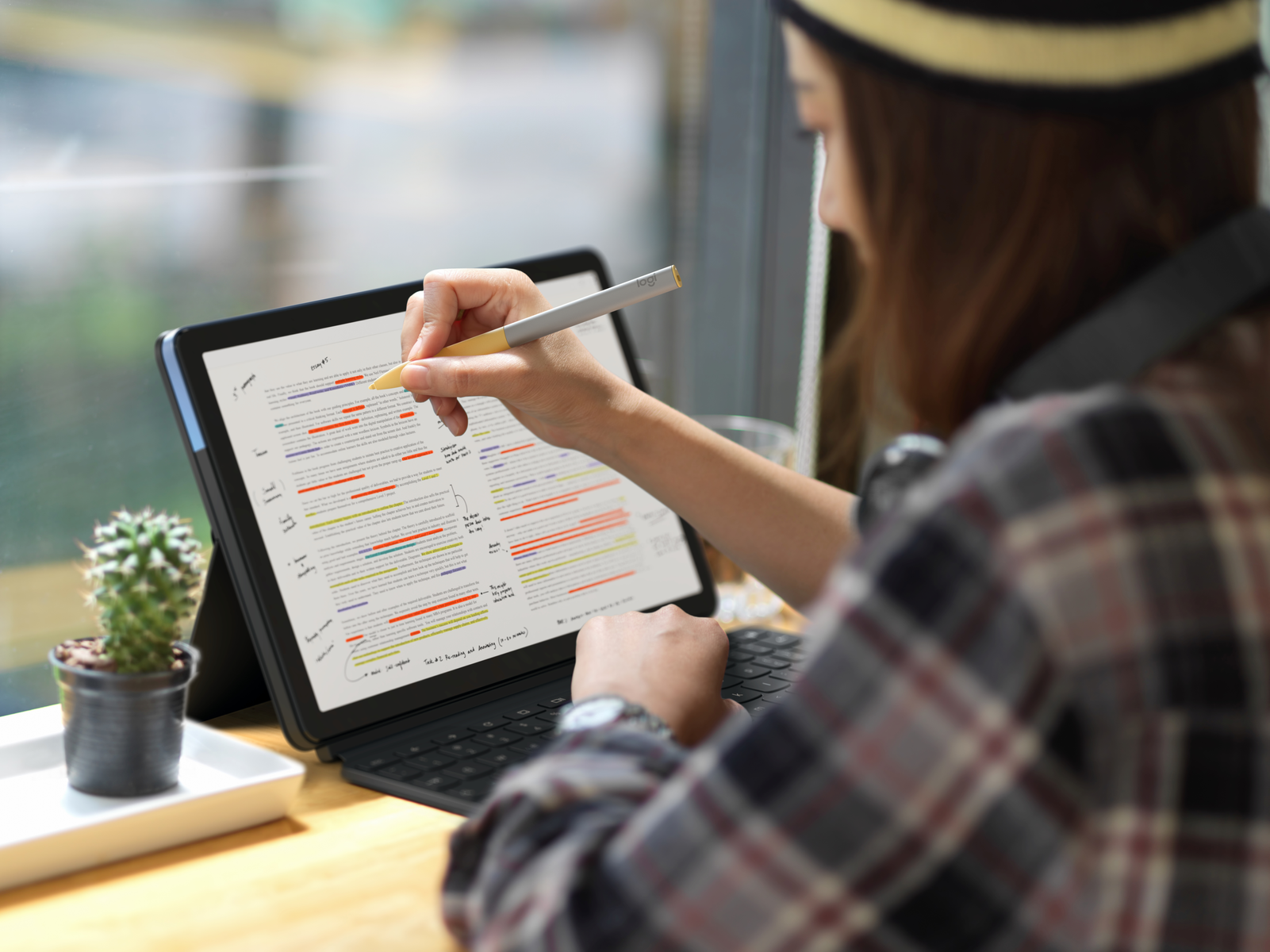 Osoba używająca pióra do zaznaczania na tablecie. Widoczny stół z rośliną.