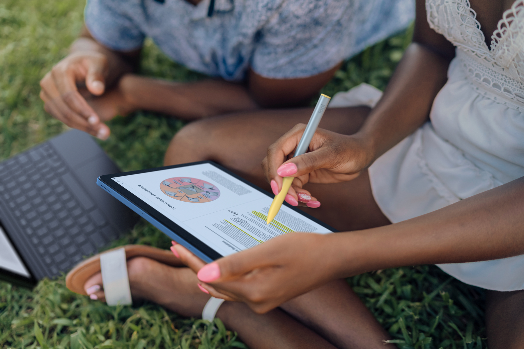 Dwie osoby siedzące na trawie patrzą na tablet. Jedna używa rysika do podświetlania tekstu.