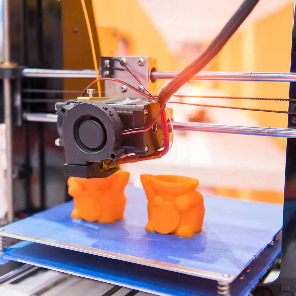 Ein 3D-Drucker druckt orangefarbene Objekte. Zwei orangefarbene Objekte auf einer blauen Oberfläche. Maschinendetail nah.