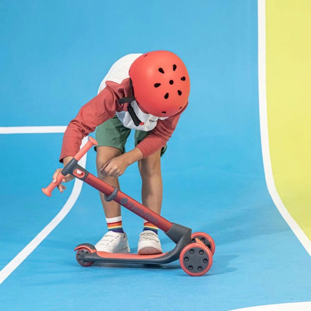 Ein Kind mit Helm steht neben einem Roller. Der Roller ist rot und grau.