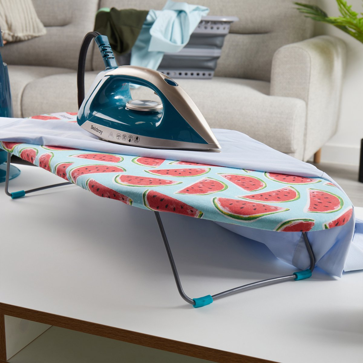 Table à repasser avec un fer teal dessus. La table a un motif de pastèque. Une chemise bleue est repassée.