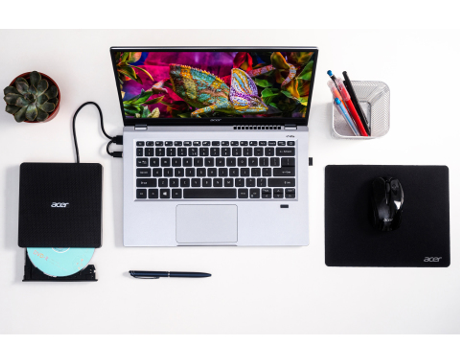Laptop z kolorowym ekranem, myszą i napędem DVD na białym biurku. Obecne są również długopisy i roślina.