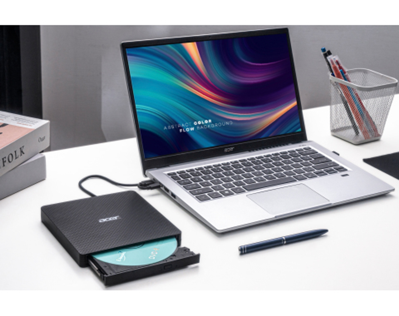 Laptop wyświetlający kolorowy abstrakcyjny obraz, z zewnętrznym napędem CD, długopisem i książkami na białym biurku.