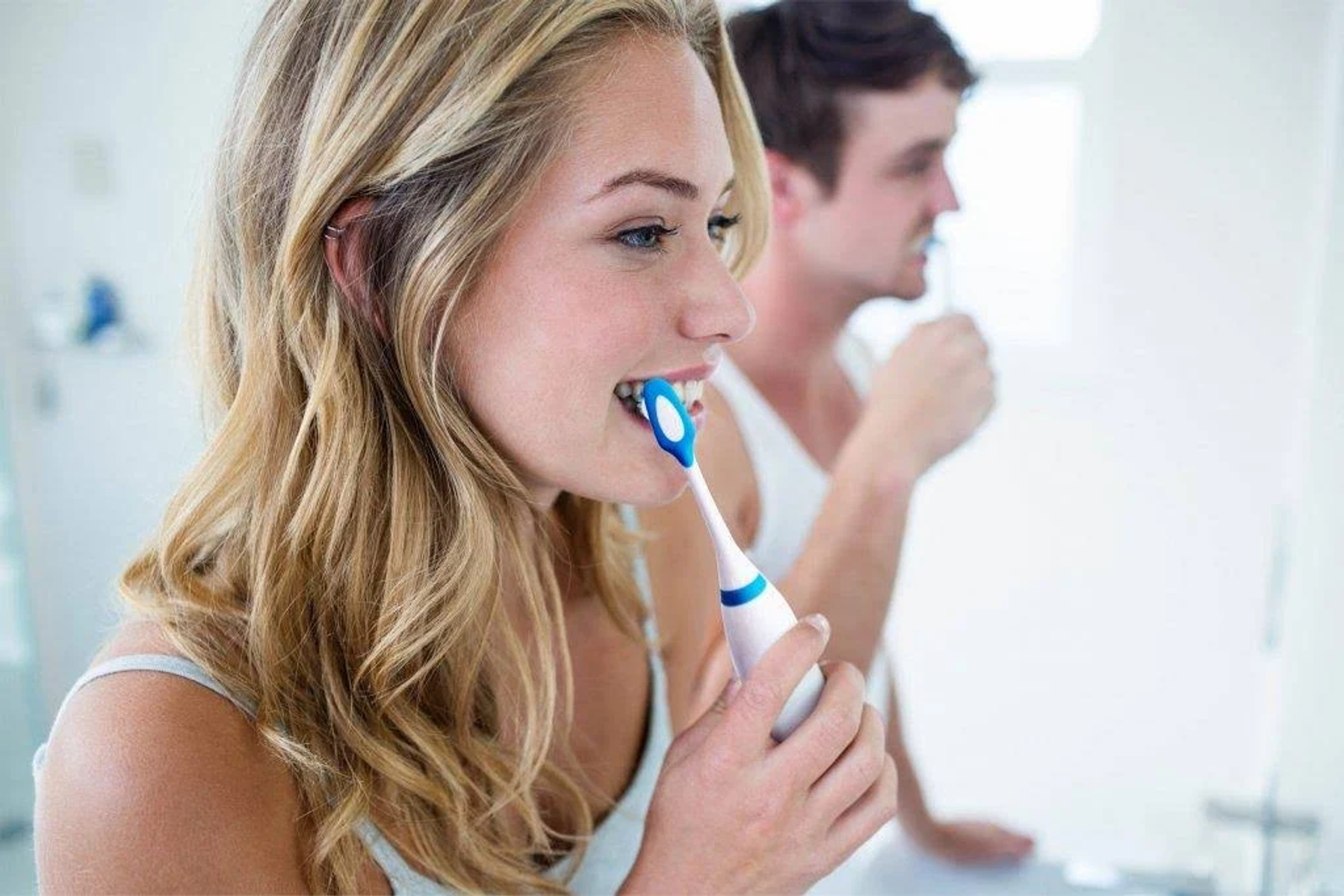 Donna che si lava i denti con spazzolino elettrico. Uomo sullo sfondo che si lava i denti.