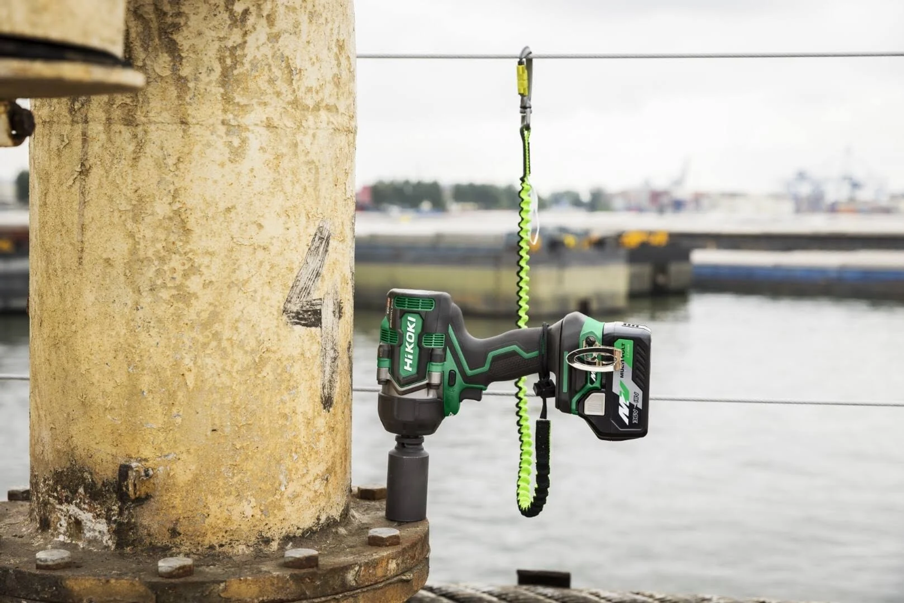 Trapano avvitatore verde e nero appeso a un palo di metallo. Sfondo: acqua, edifici e cielo nuvoloso.