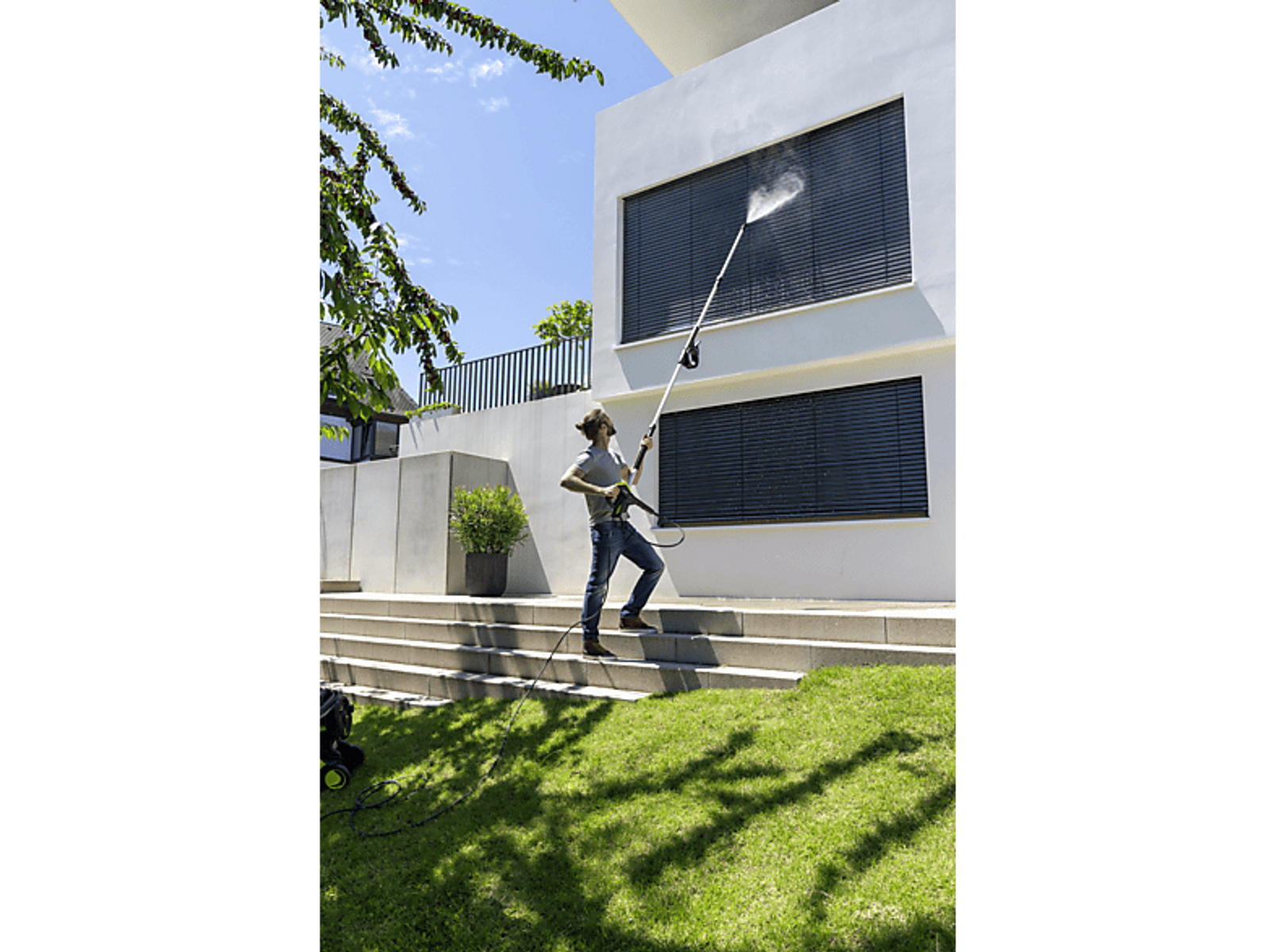 Hombre usando una hidrolavadora en una casa. Césped verde, casa blanca y cielo azul al fondo.