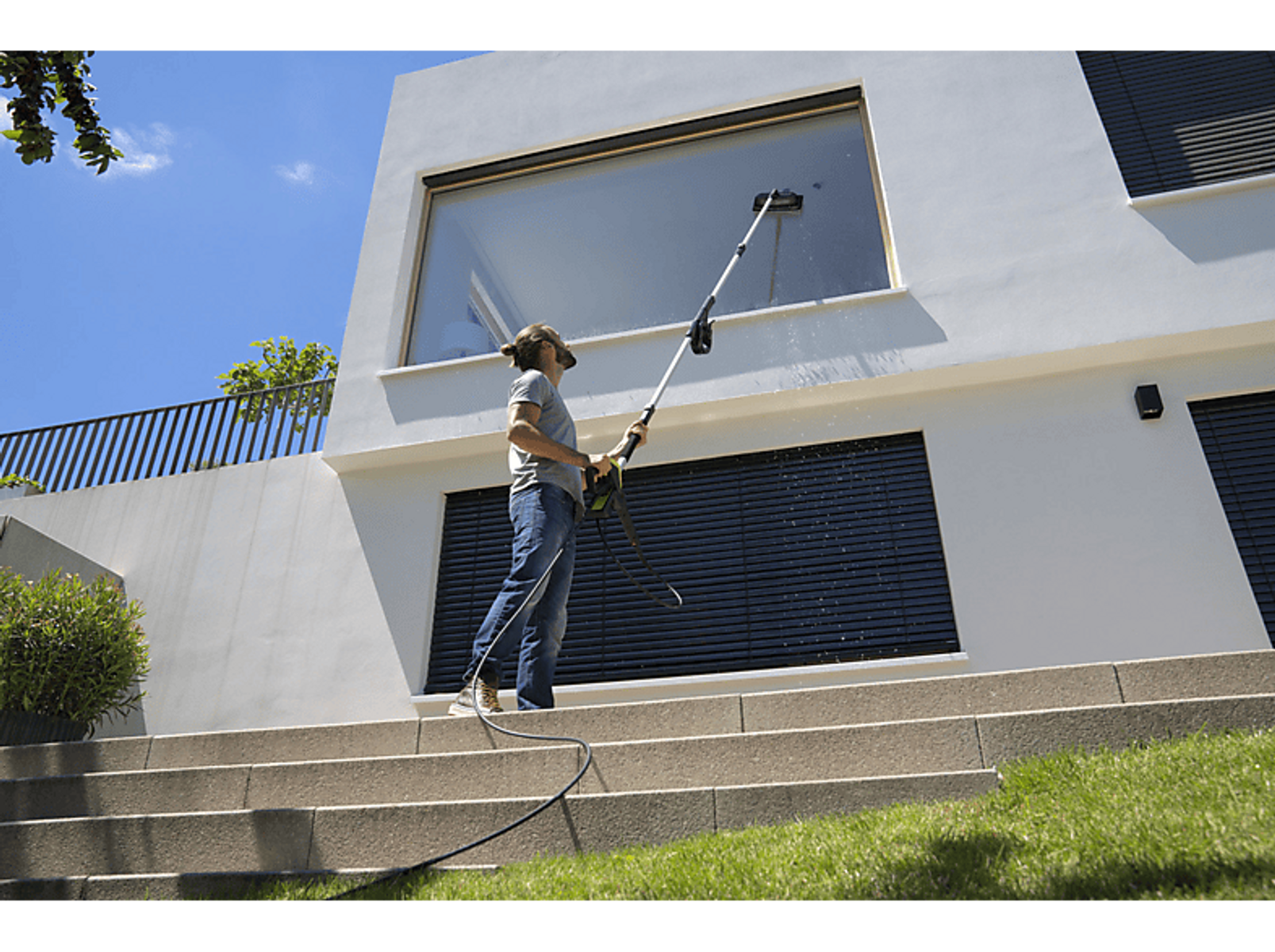 Hombre limpiando una ventana en una casa con un poste largo. Cielo azul, hierba verde y escalones de piedra.