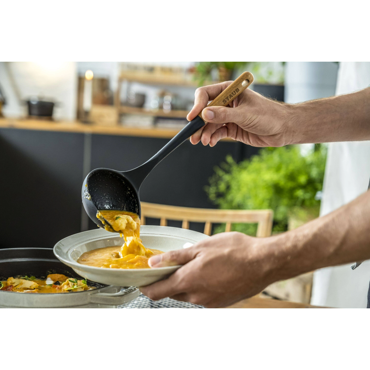 Hände gießen Suppe aus einer schwarzen Schöpfkelle in eine weiße Schüssel. Etwas Essen in der Pfanne links.