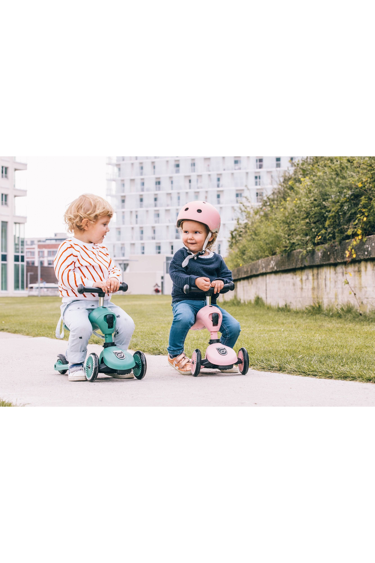 Dwoje dzieci na hulajnogach. Jedno ma kask. Tło: budynki i zieleń.