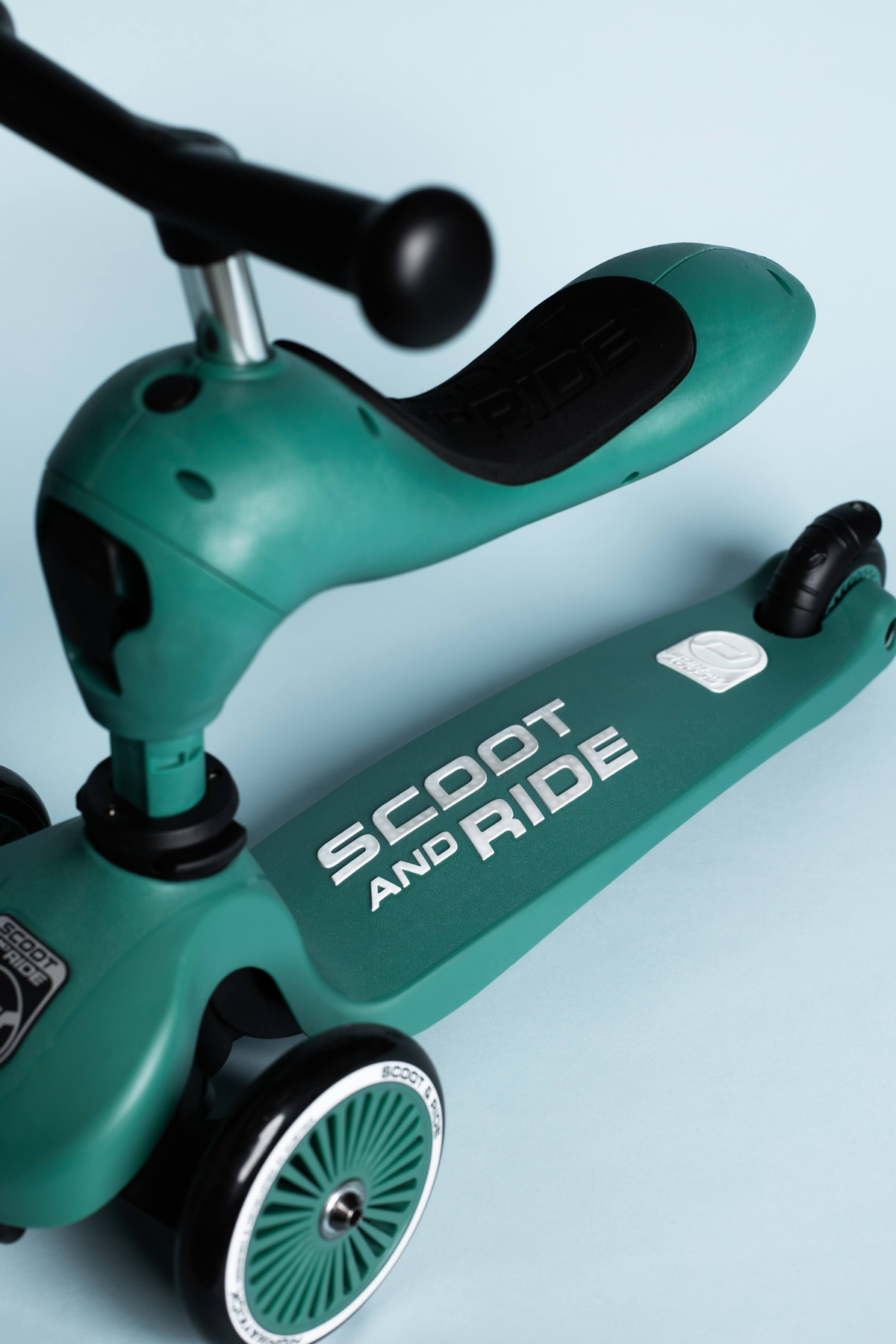 Zbliżenie zielonej hulajnogi. Ukazuje siedzenie, koła i logo 'SCOOT AND RIDE'.