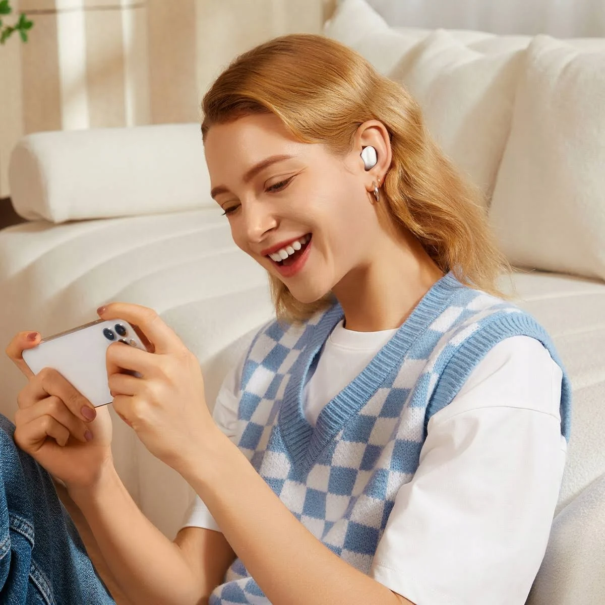 Una donna sorridente, con auricolari e che usa uno smartphone. È seduta su un divano.