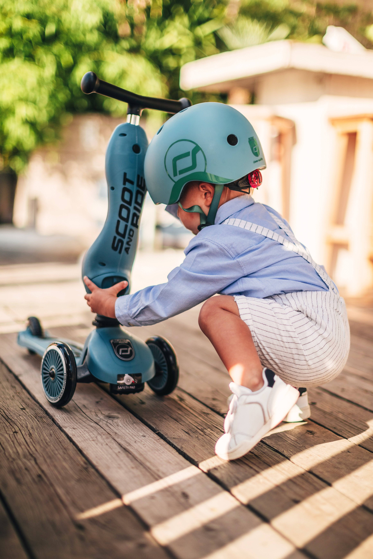 Bambino con casco e scooter su piattaforma in legno. Il bambino è accovacciato vicino allo scooter.