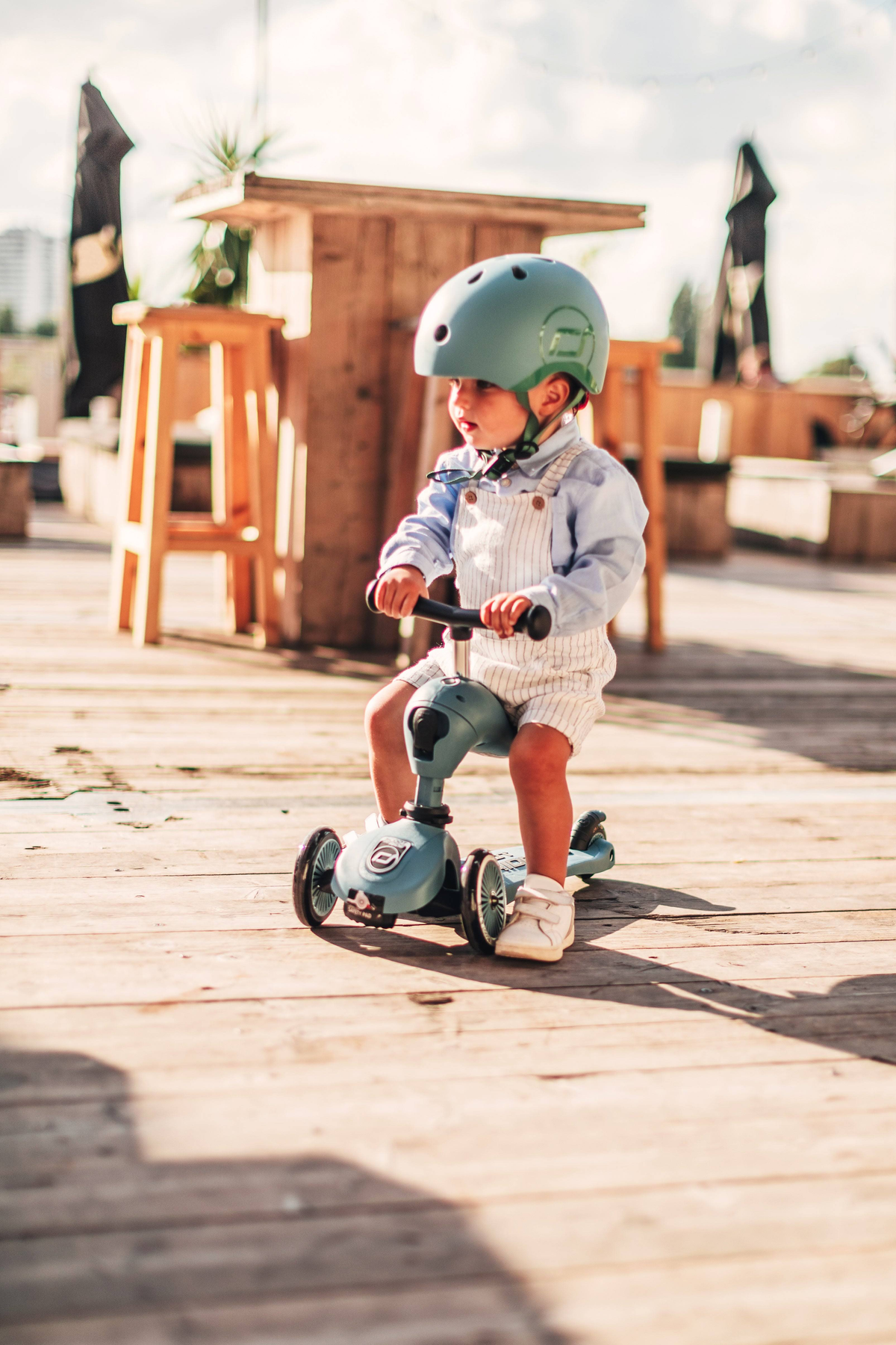 Bambino su scooter blu su piattaforma in legno, con casco. Ambiente esterno soleggiato.