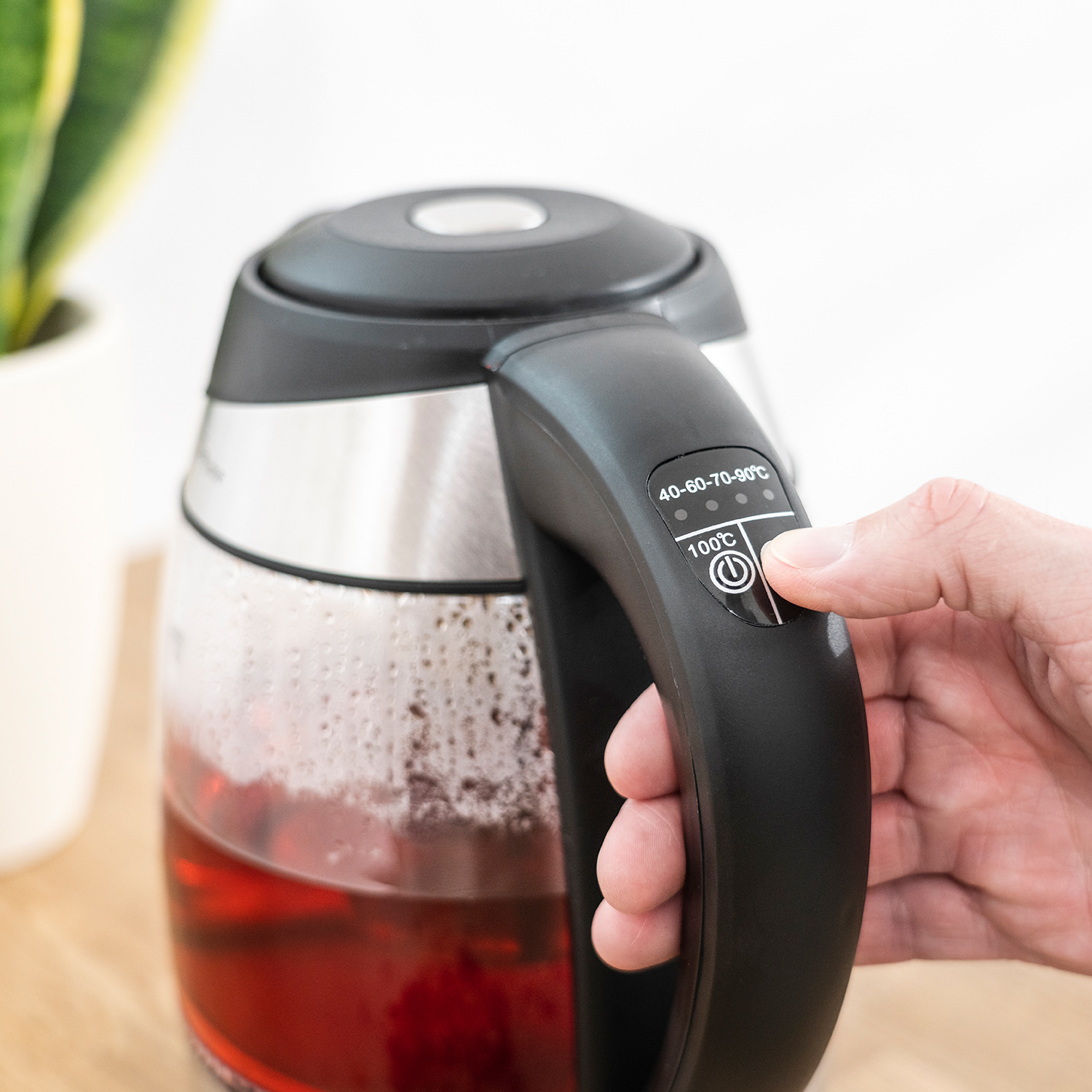 Elektrischer Wasserkocher mit einer Hand, die einen Knopf drückt. Anzeige zeigt Temperatureinstellungen. Tee im Wasserkocher.