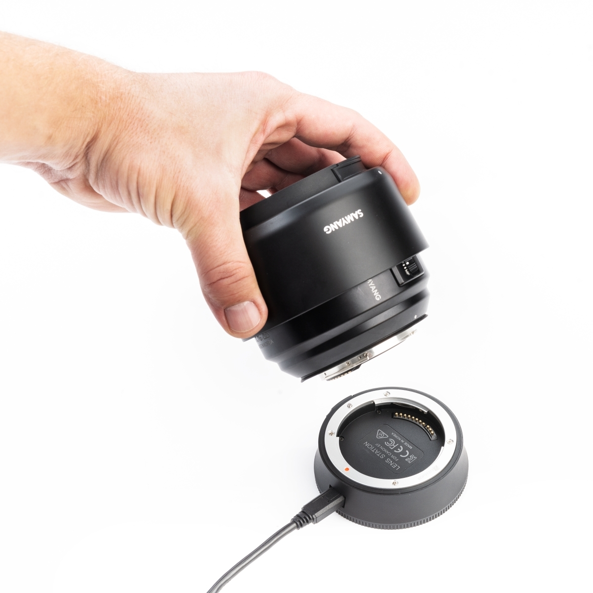 Eine Hand hält ein schwarzes Objektiv über einer schwarzen Objektivstation mit USB-Kabel.