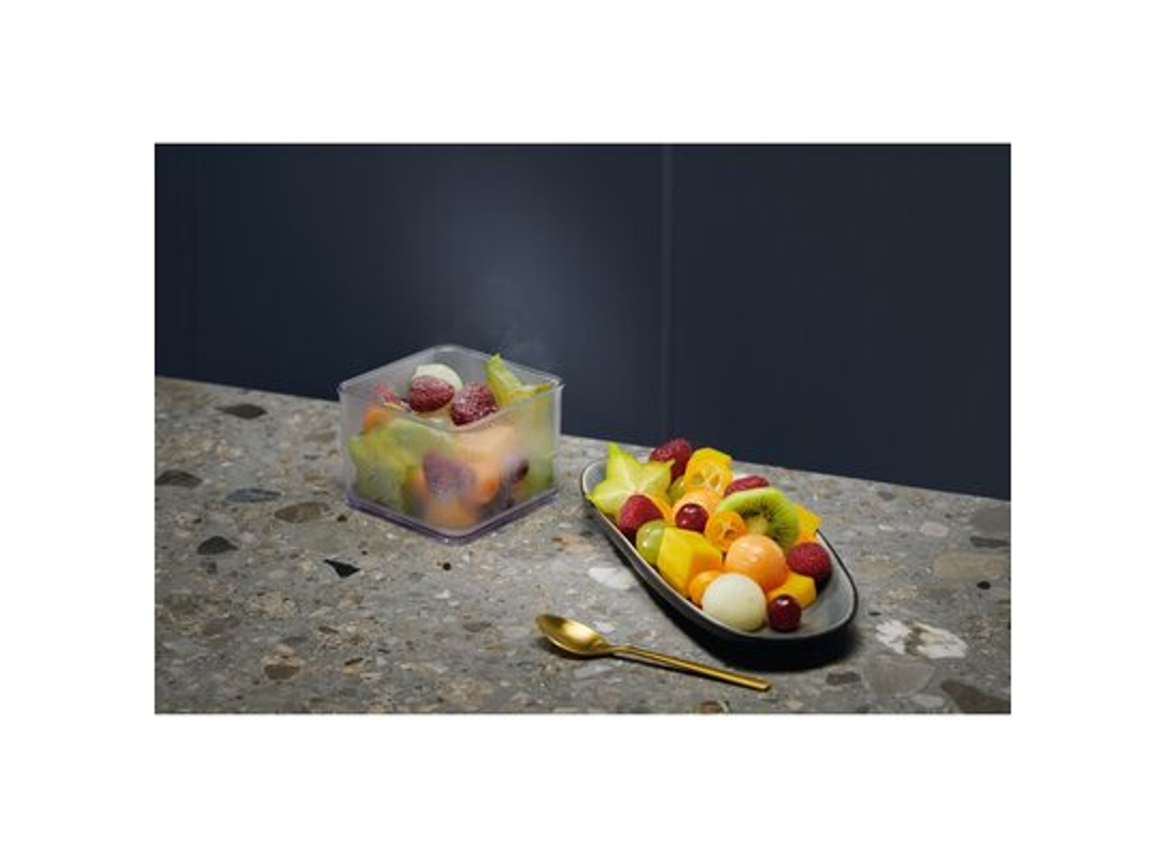 Un cuenco de ensalada de frutas y un recipiente de fruta congelada, con una cuchara dorada sobre una superficie gris.