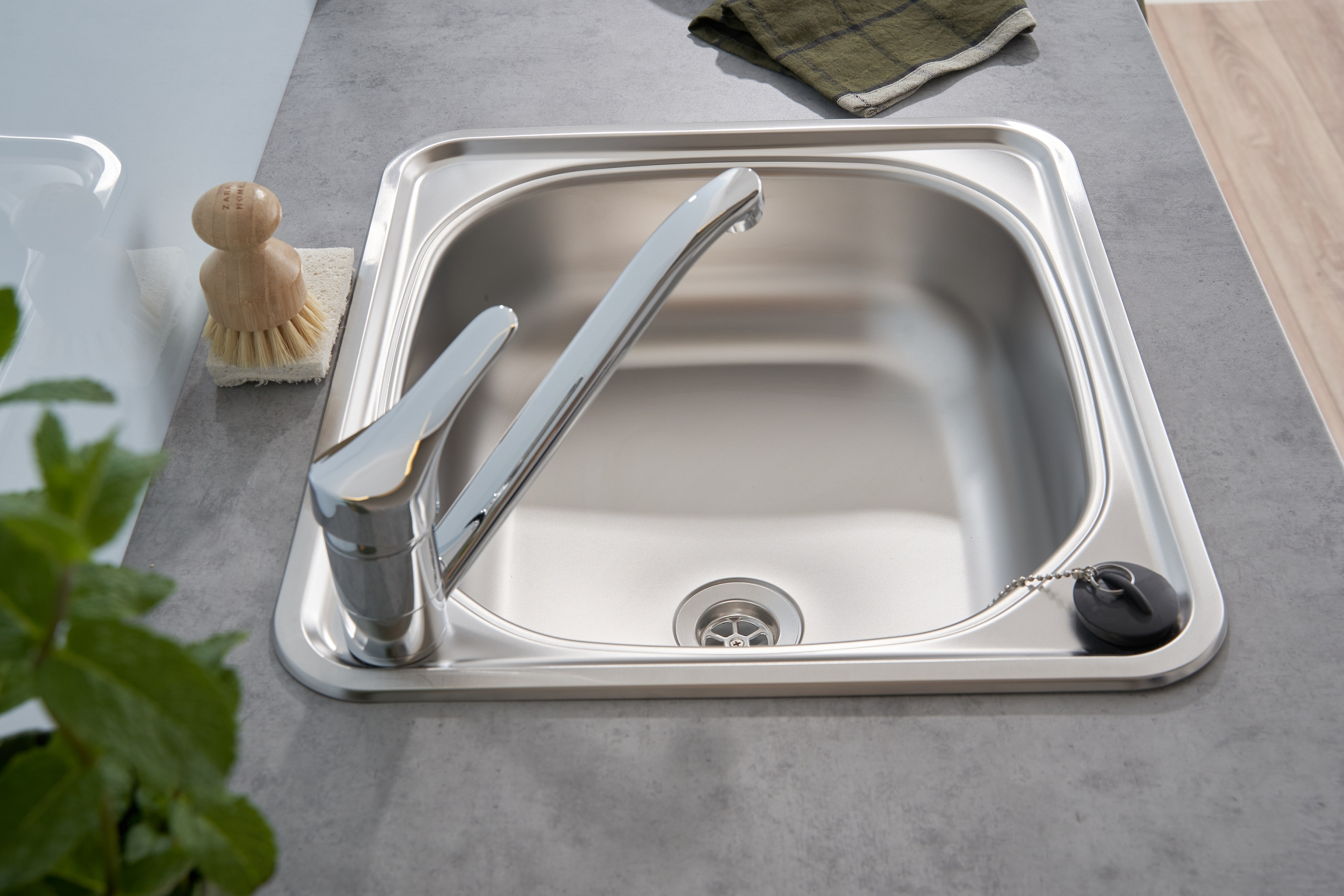 Edelstahl-Küchenspüle mit Wasserhahn, Abfluss und Bürste, auf einer grauen Arbeitsplatte, Draufsicht.