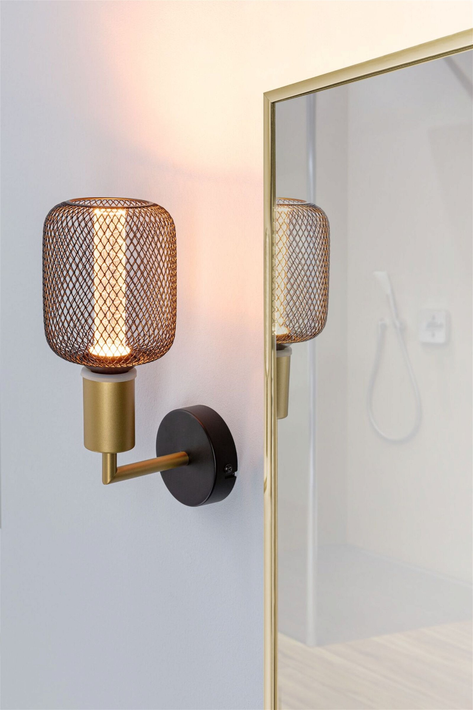 Lampa ścienna z czarnym metalowym kloszem z siatki, złotym ramieniem i odbiciem w lustrze.