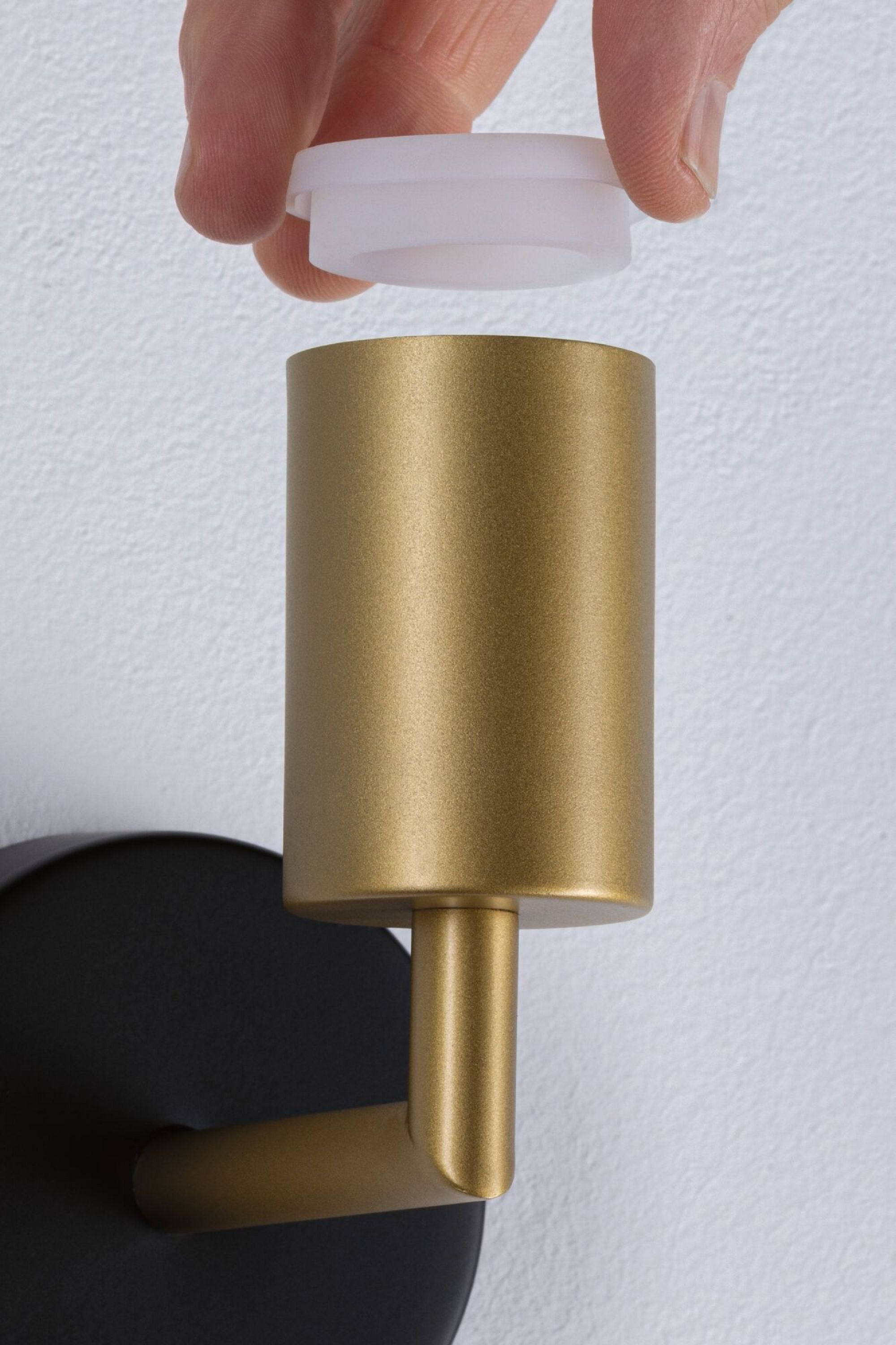 Ręka umieszcza białą nasadkę na złotej, cylindrycznej lampie ściennej.