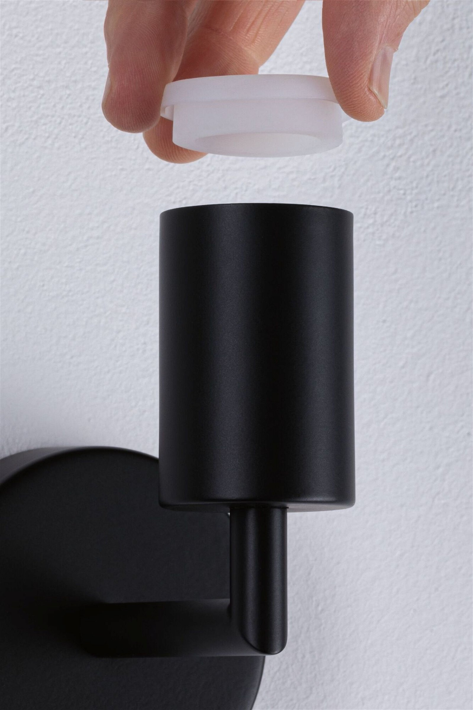 Dłoń umieszczająca białą nasadkę na czarnej lampie na białej ścianie.