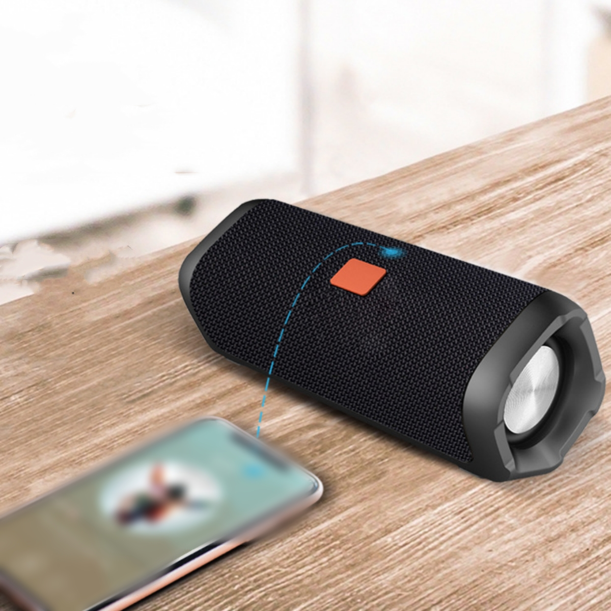 Schwarz-silberner Lautsprecher auf Holztisch mit verschwommenem Smartphone davor, orangefarbener Knopf oben.
