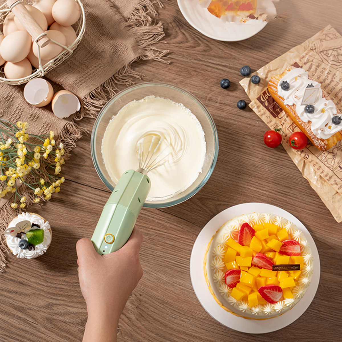 Eine Hand verwendet einen Mixer, um Sahne in einer Glasschüssel zu schlagen. Kuchen und Eier sind ebenfalls zu sehen.