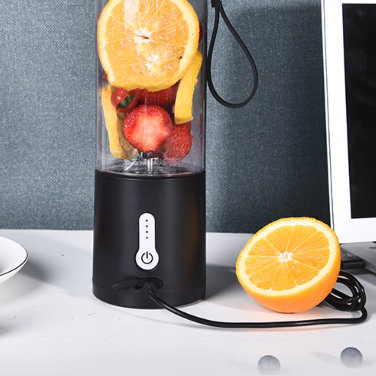 Ein schwarzer Mixer mit Obst im Inneren, eine halbe Orange daneben und ein Computer im Hintergrund.