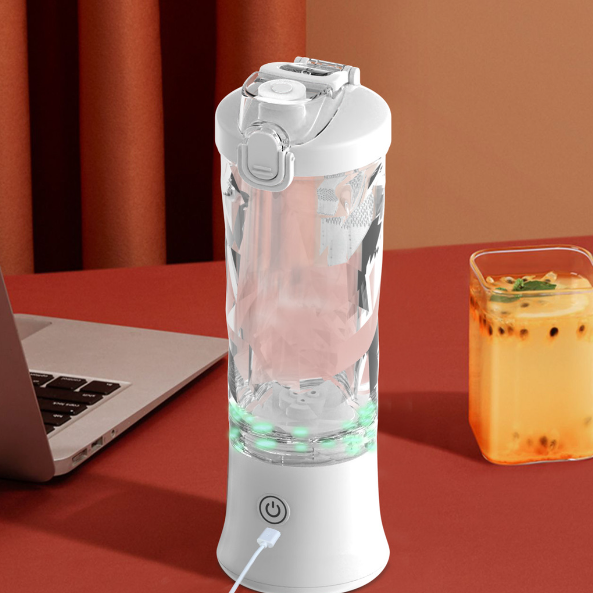 Ein weißer tragbarer Mixer mit einem transparenten Behälter, Ladekabel und einem Getränk im Hintergrund.