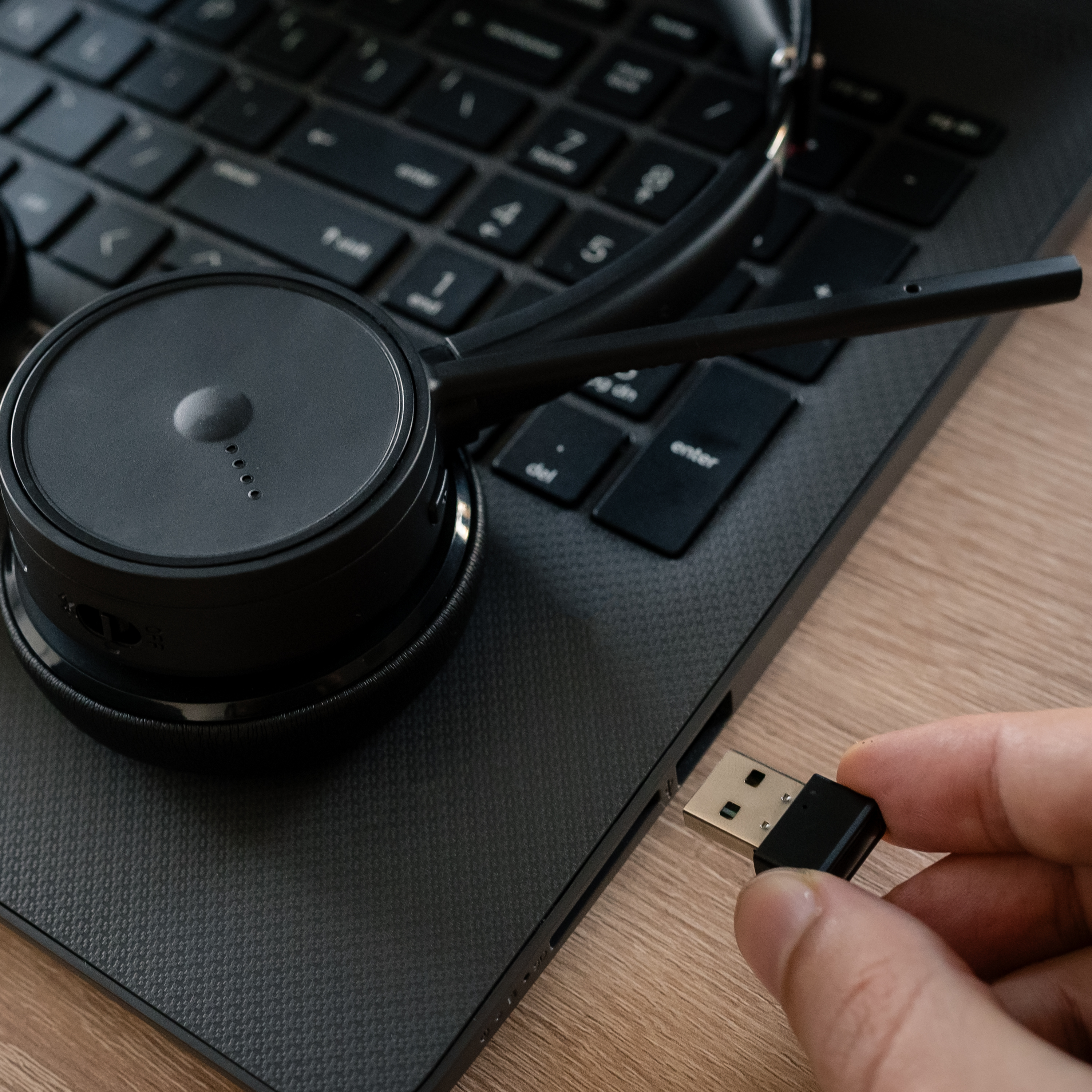 Die Hand einer Person, die einen schwarzen USB-Adapter in einen Laptop steckt, mit einem Headset in der Nähe.