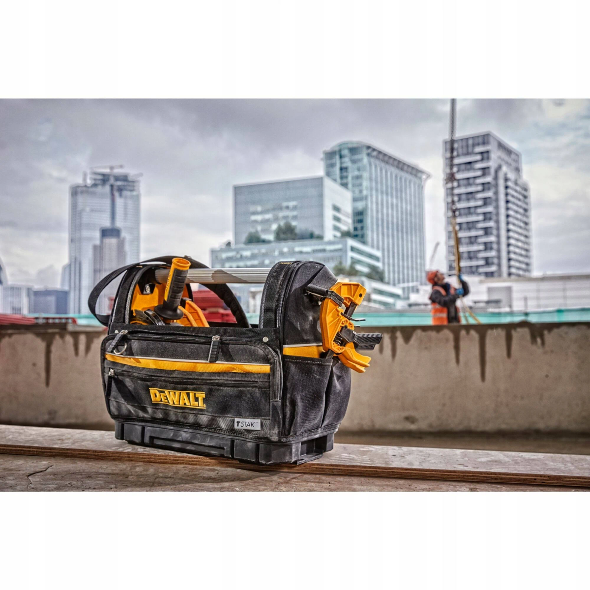 Offene schwarz-gelbe Werkzeugtasche auf einer Baustelle mit Gebäuden im Hintergrund.
