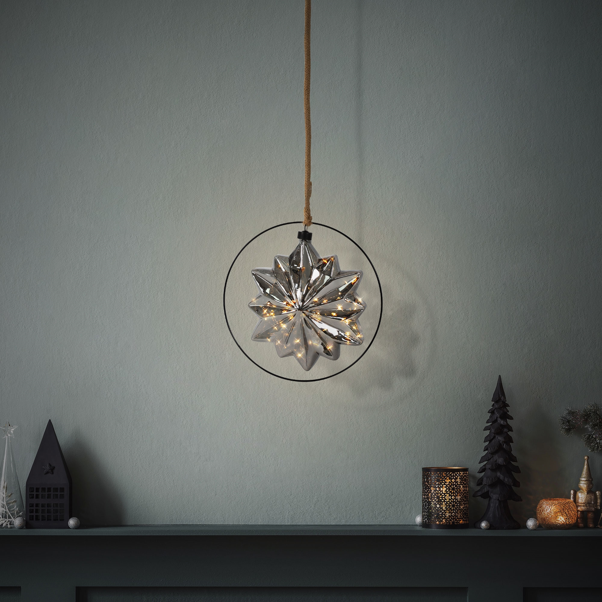 Ein silberner, sternförmiger Schmuck mit warmweißen Lichtern hängt. Ein Regal mit Dekorationen an einer dunkelgrünen Wand.