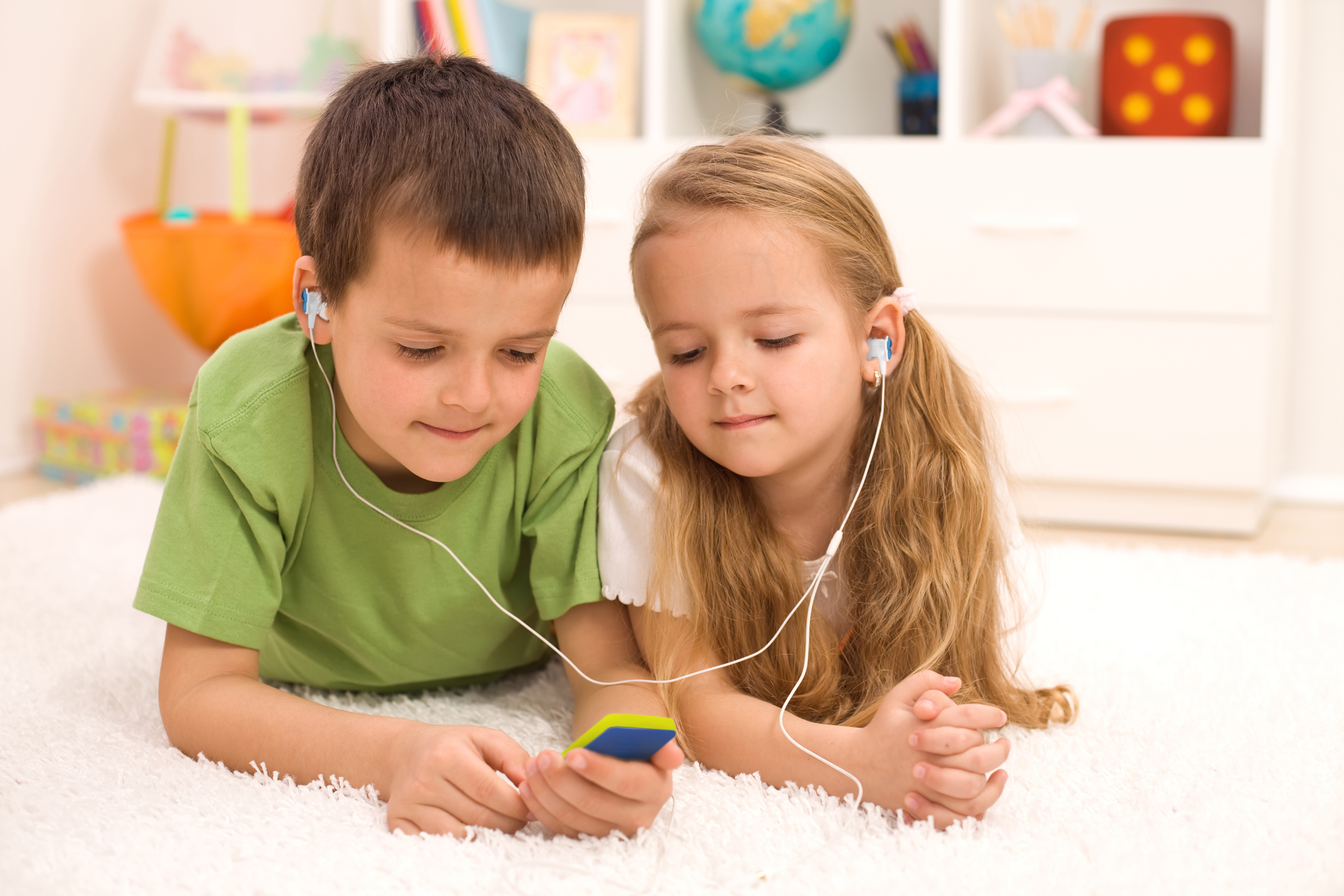 Zwei Kinder liegen auf einem Teppich und hören Musik auf einem Gerät mit Kopfhörern.