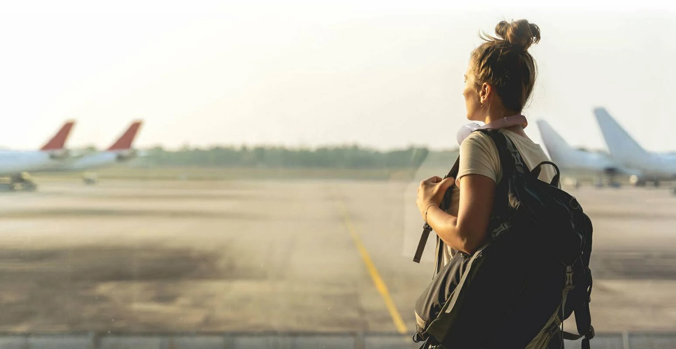 Frau schaut durch ein Glasfenster am Flughafen auf Flugzeuge, mit einem Rucksack.