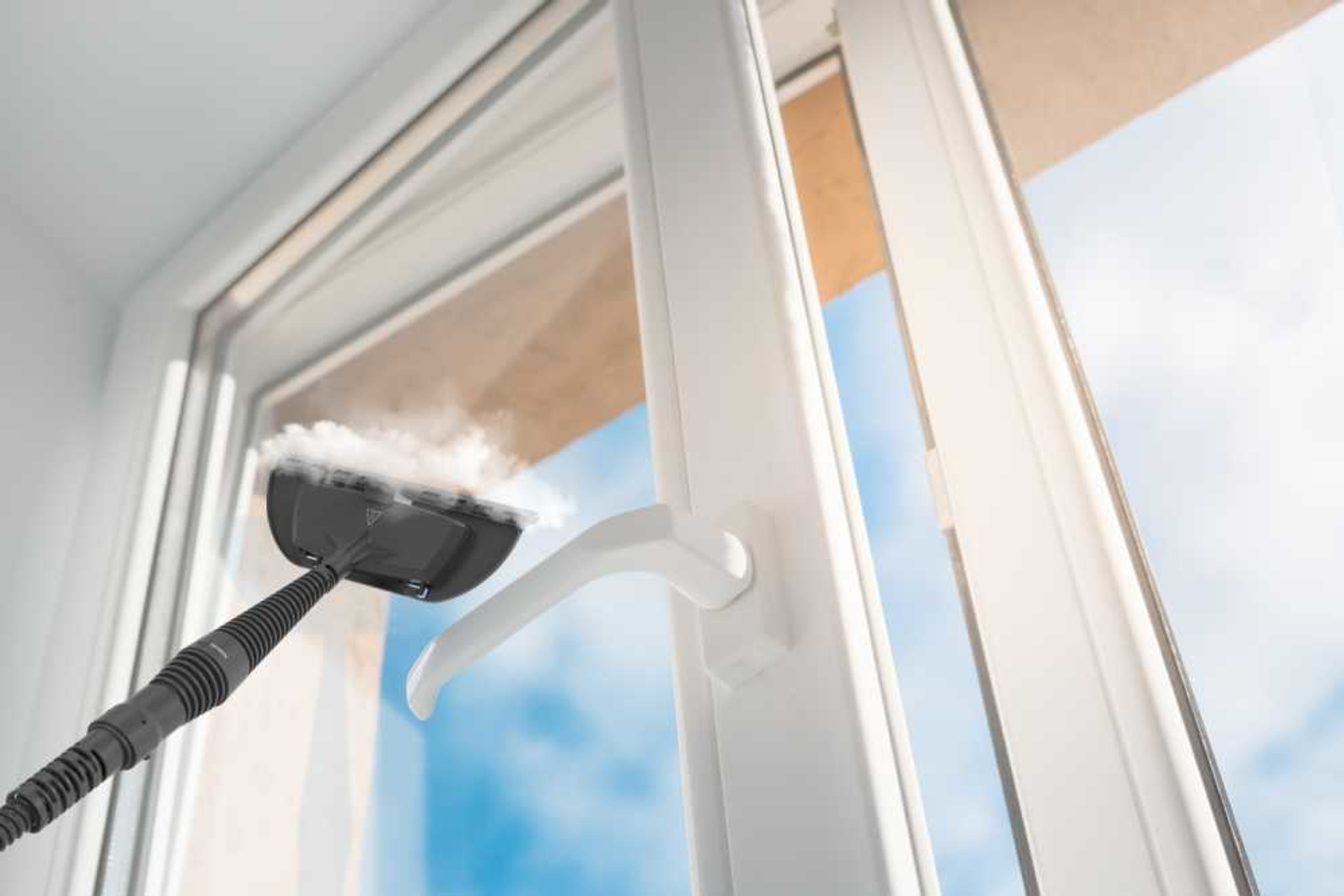 Oczyszczacz parowy czyszczący okno. Biała rama, widoczne niebieskie niebo. Para wydobywa się z oczyszczacza.