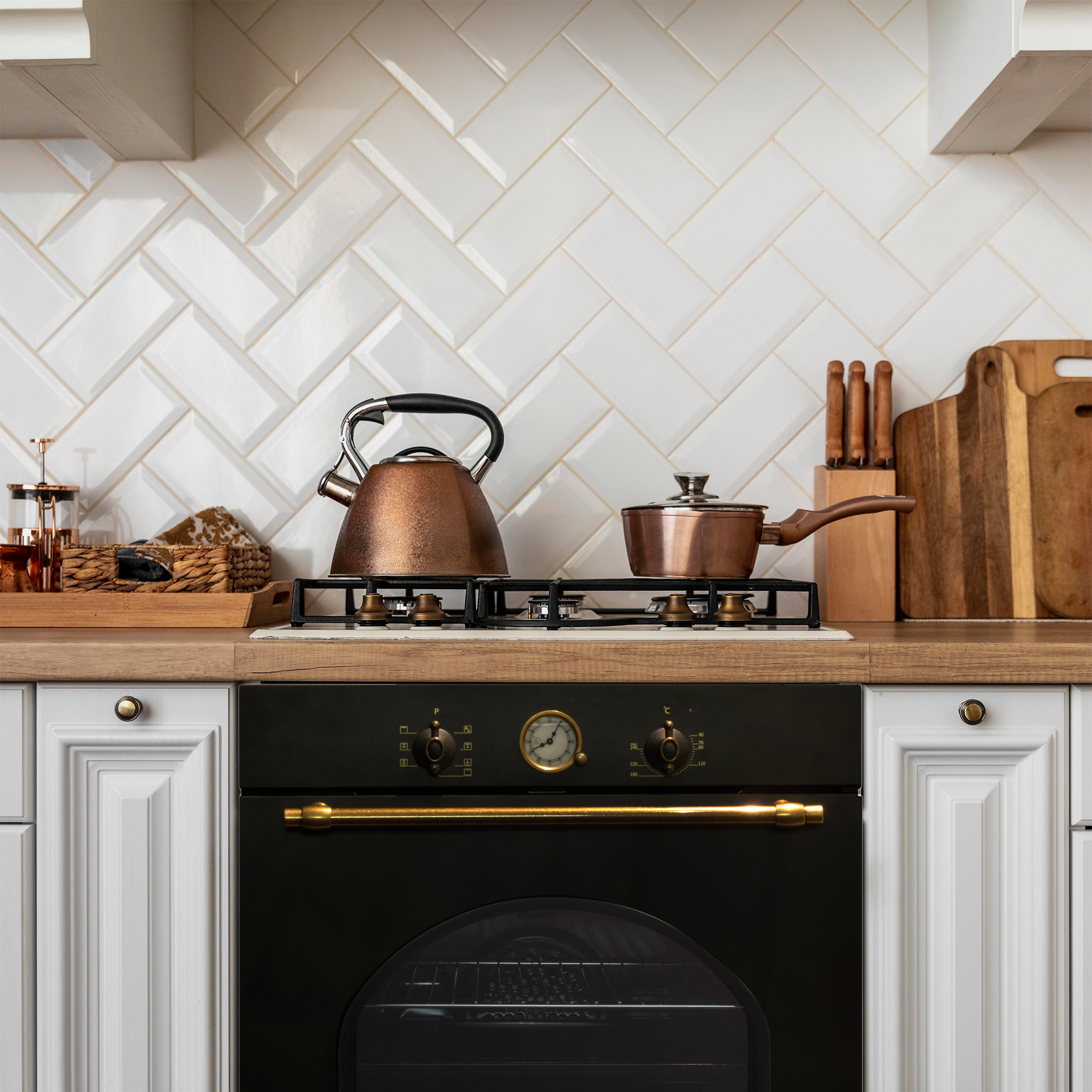 Kuchnia z kuchenką, miedzianym czajnikiem i garnkiem. Biała ściana z płytek i drewniane blaty.