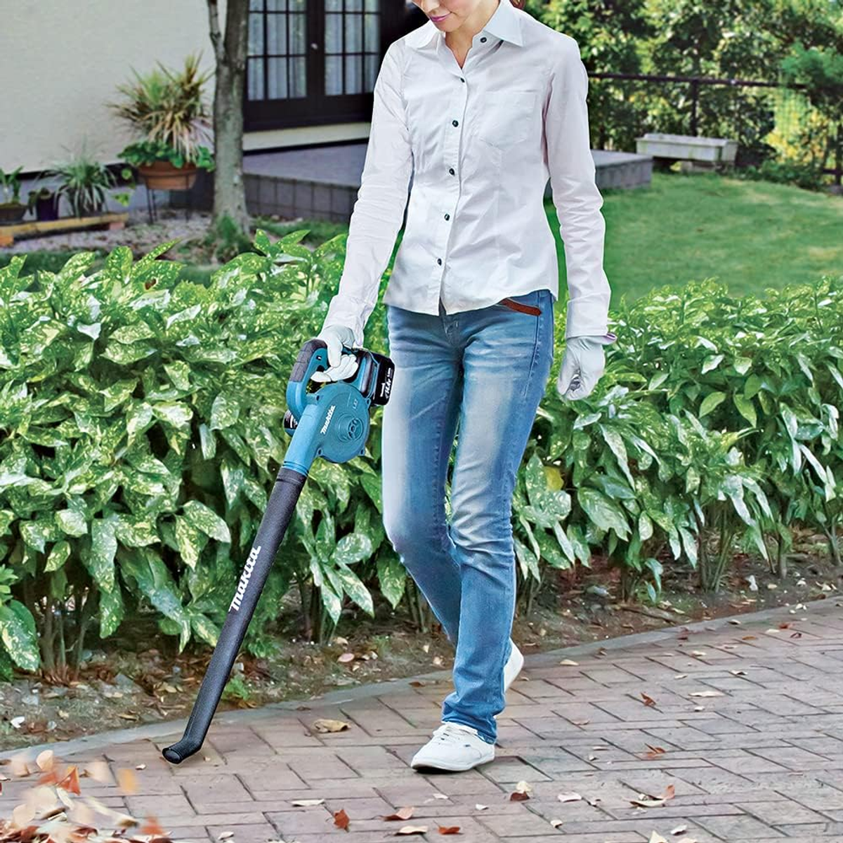 A woman using a leaf blower to clean a brick path. She wears a white shirt and blue jeans.