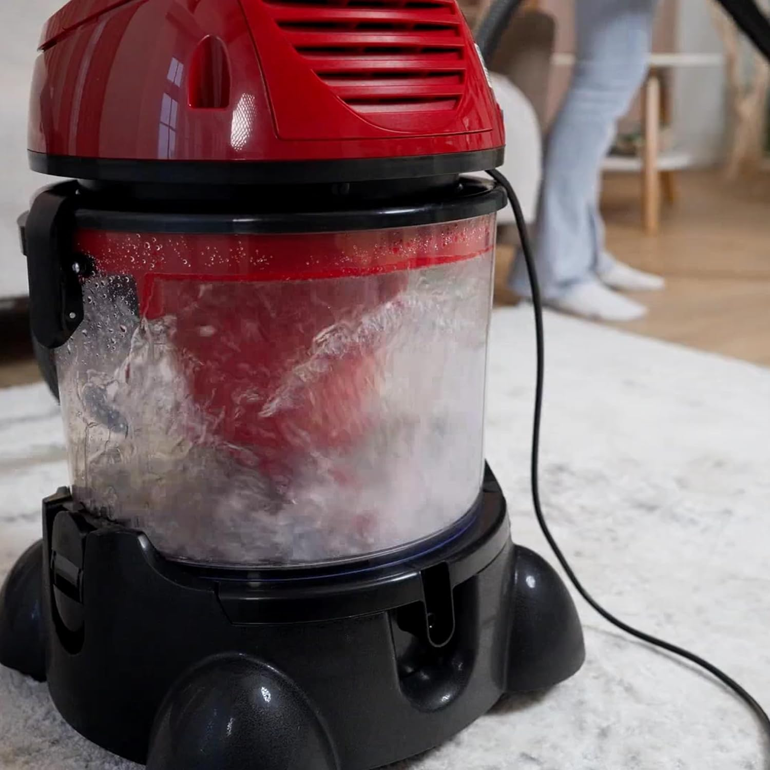 Roter und schwarzer Nass-Trocken-Staubsauger mit Wasser, auf einem Teppich, mit Person im Hintergrund.