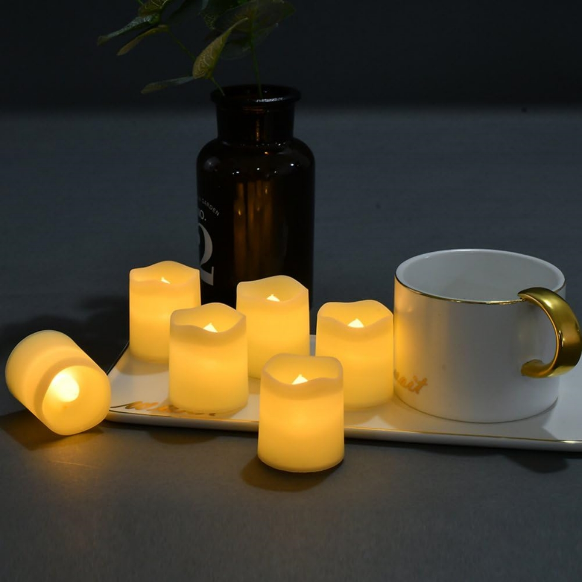 Sechs kleine, gelbe LED-Kerzen mit einer weißen Tasse und einer schwarzen Vase. Alle Objekte sind auf einem weißen Tablett.