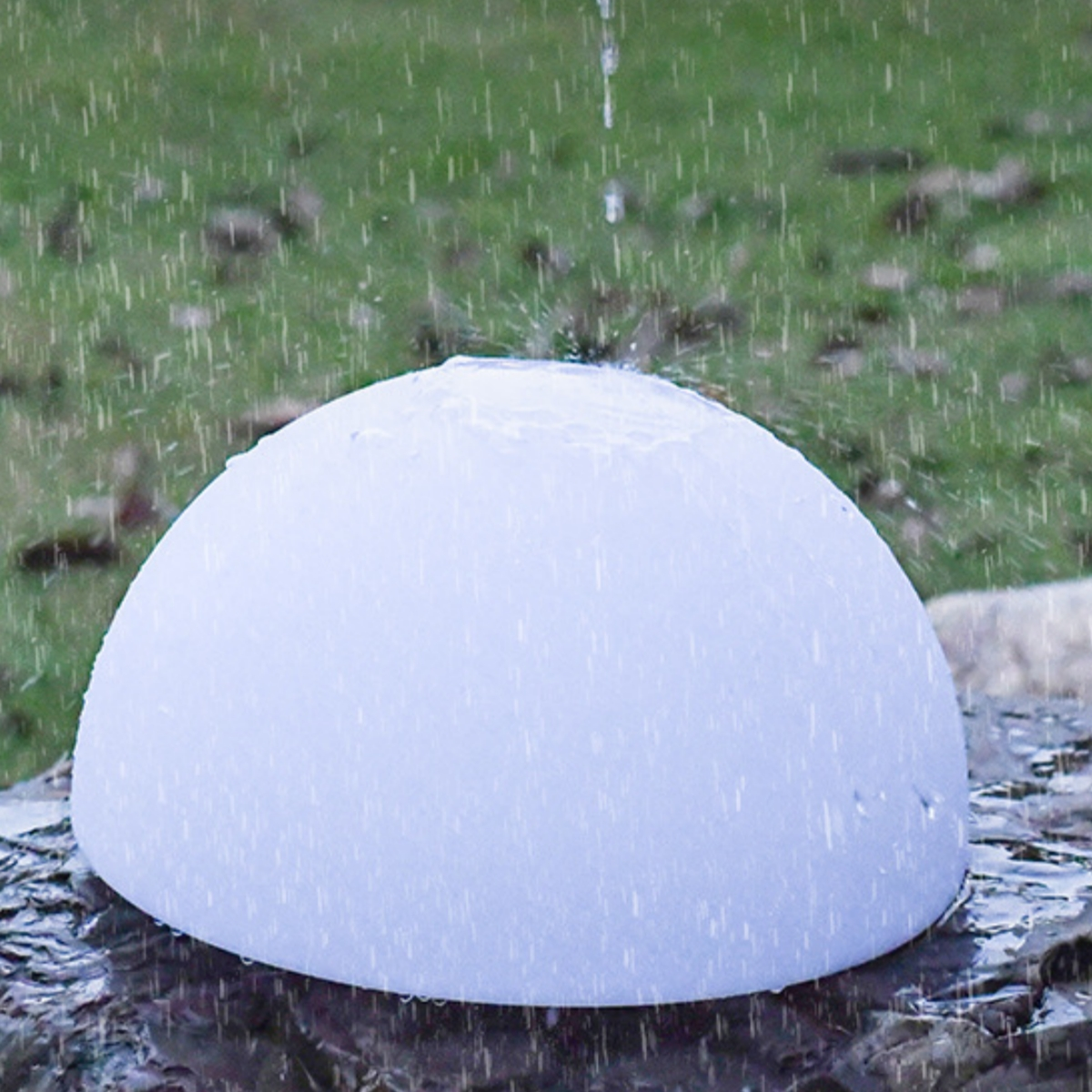 Ein weißes, kuppelförmiges Objekt steht auf einem Felsen, Regen fällt. Der Hintergrund ist verschwommenes grünes Gras.
