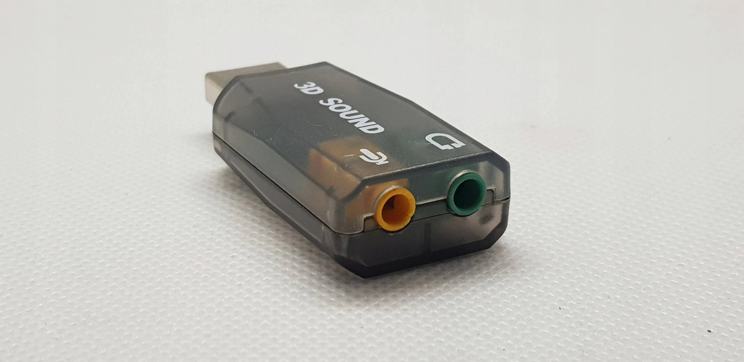 Adapter dźwięku 3D ze złączem USB i gniazdami wejścia/wyjścia audio. Znajduje się na białej powierzchni.